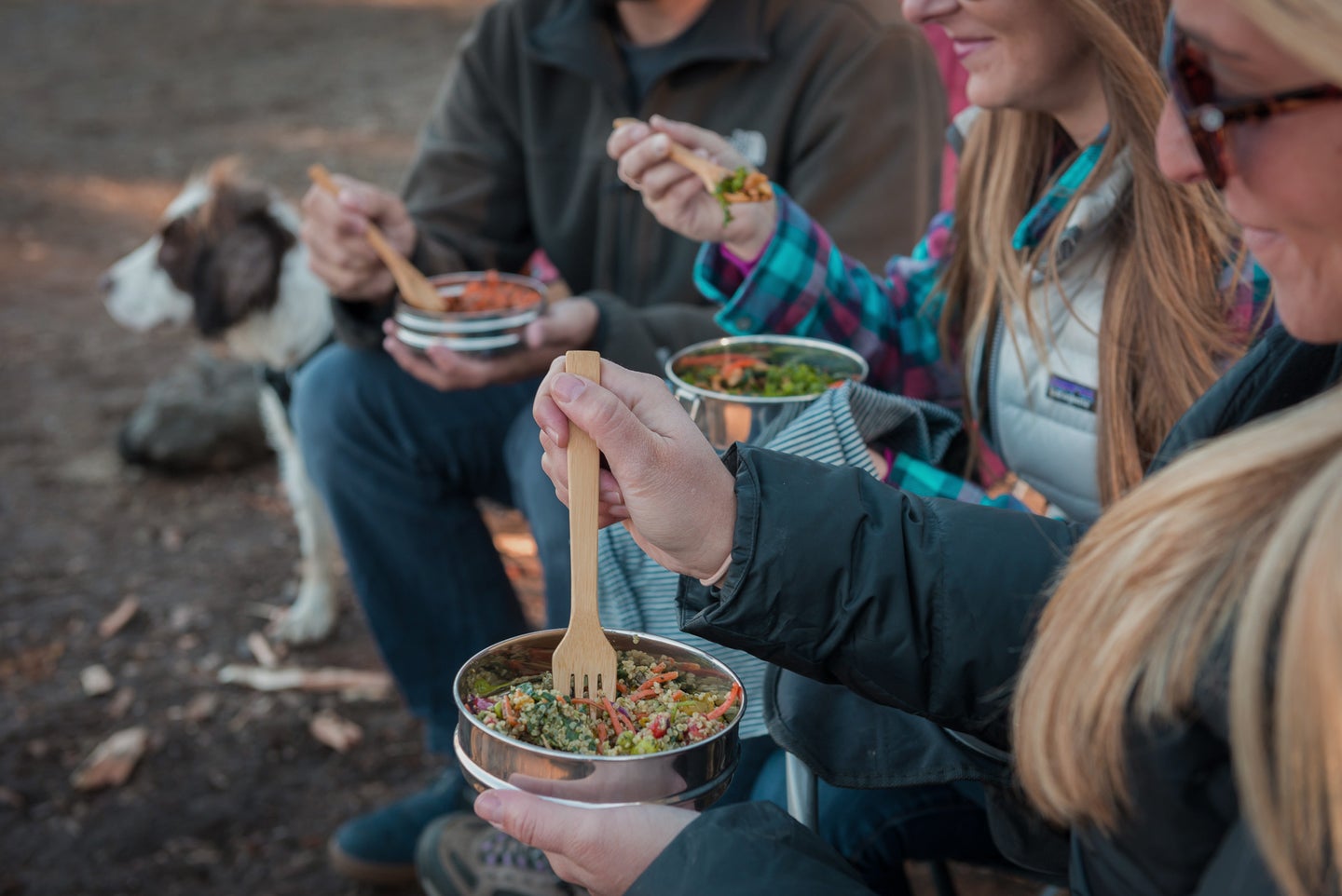 A group of people sit outside, eating a meal from stainless steel food containers using bamboo forks. A dog is in the background.