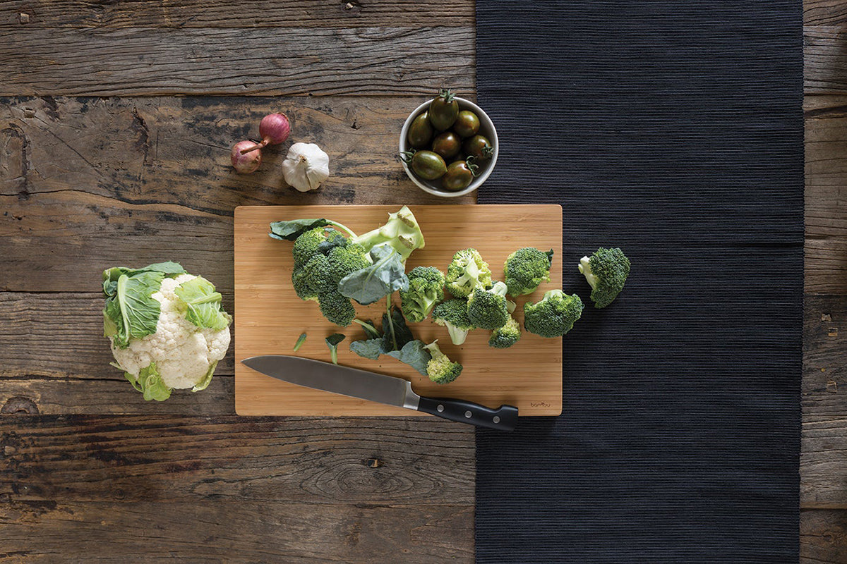 A bamboo cutting board is on a wooden tabletop. There is a head of cauliflower, some chopped broccoli, and other vegetables on and near the cutting board. A knife also rests on the cutting board.