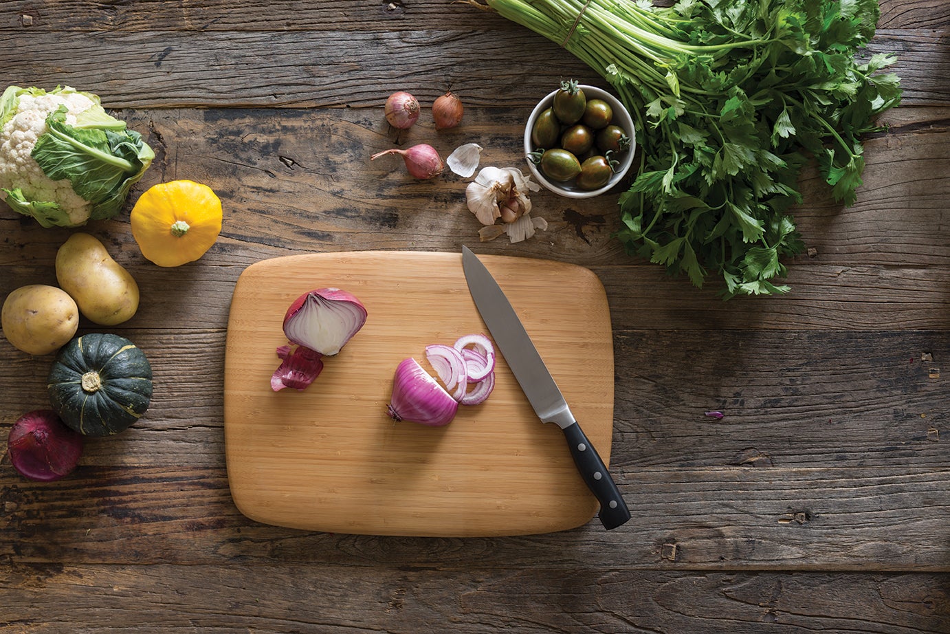 A Medium Classic Bamboo Cutting and Serving Board is shown with a knife that has been used to slice red onions. Assorted vegetables surround the cutting board.