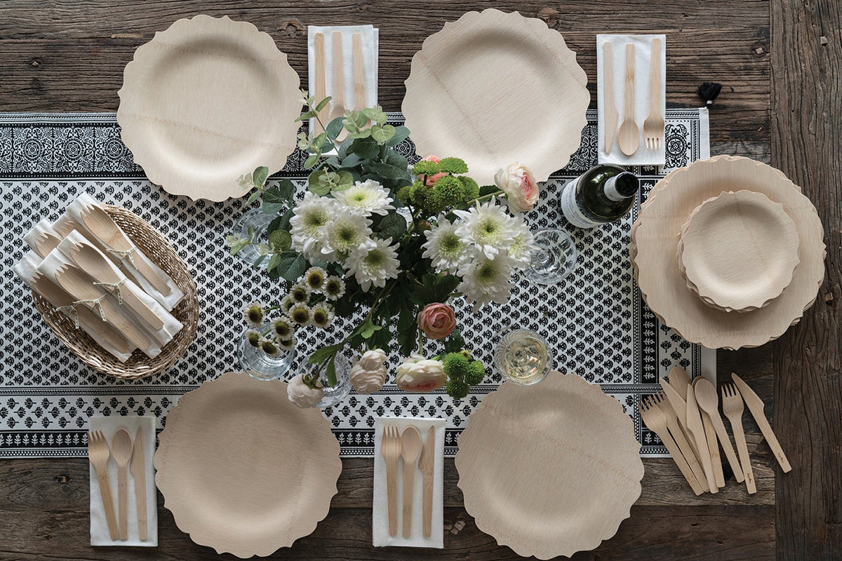 A table setting with scalloped bamboo "Fancy" plates is laid out on a blue tablecloth. There id a flower arrangement at the center of the table, and bamboo cutlery is laid out on napkins beside the plates.