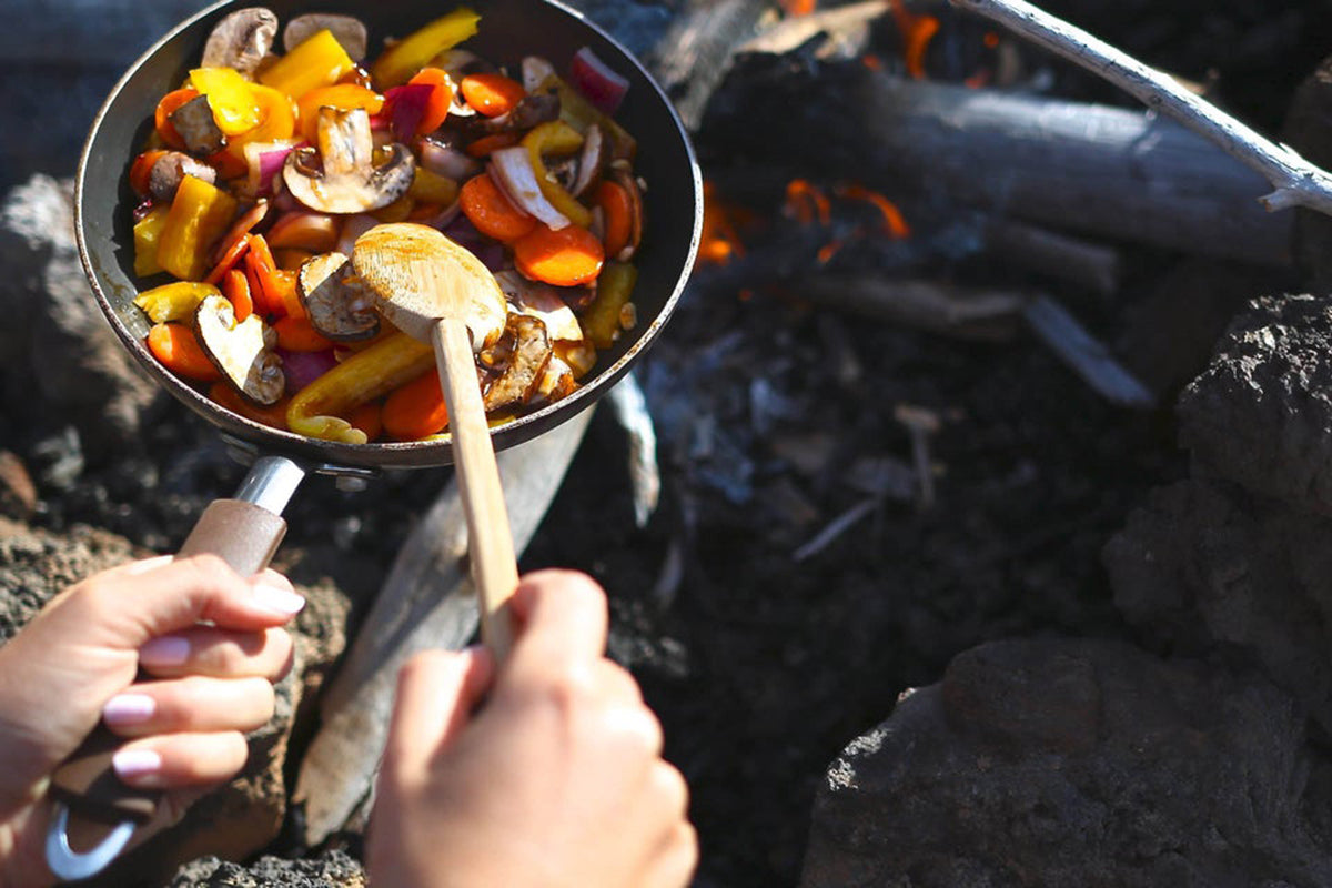 A person cooks in a skillet over a campfire. A bamboo mixing spoon is stirring vegetables as they cook.