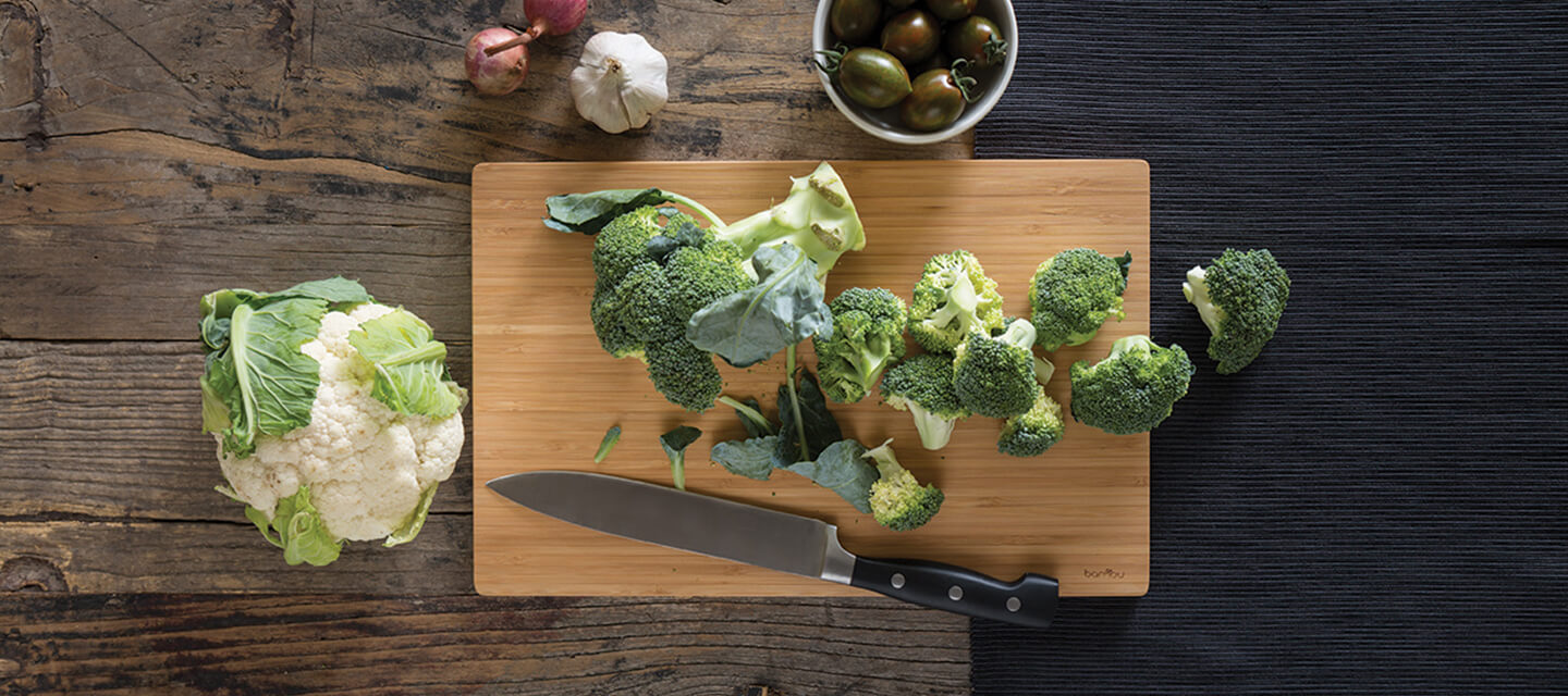 bamboo cutting board with chopped broccoli. A head of cauliflower is nearby.