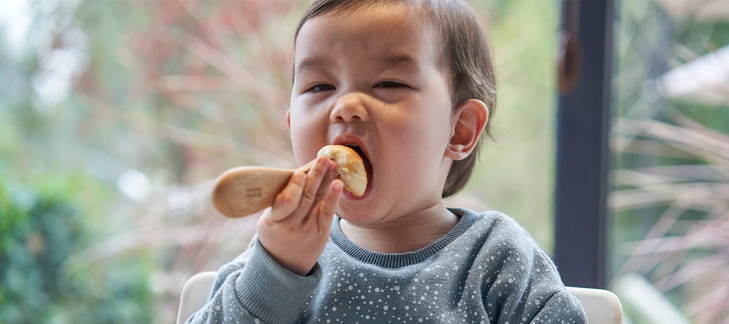 A young child in a blue sweater uses a bamboo baby utensil to eat with.