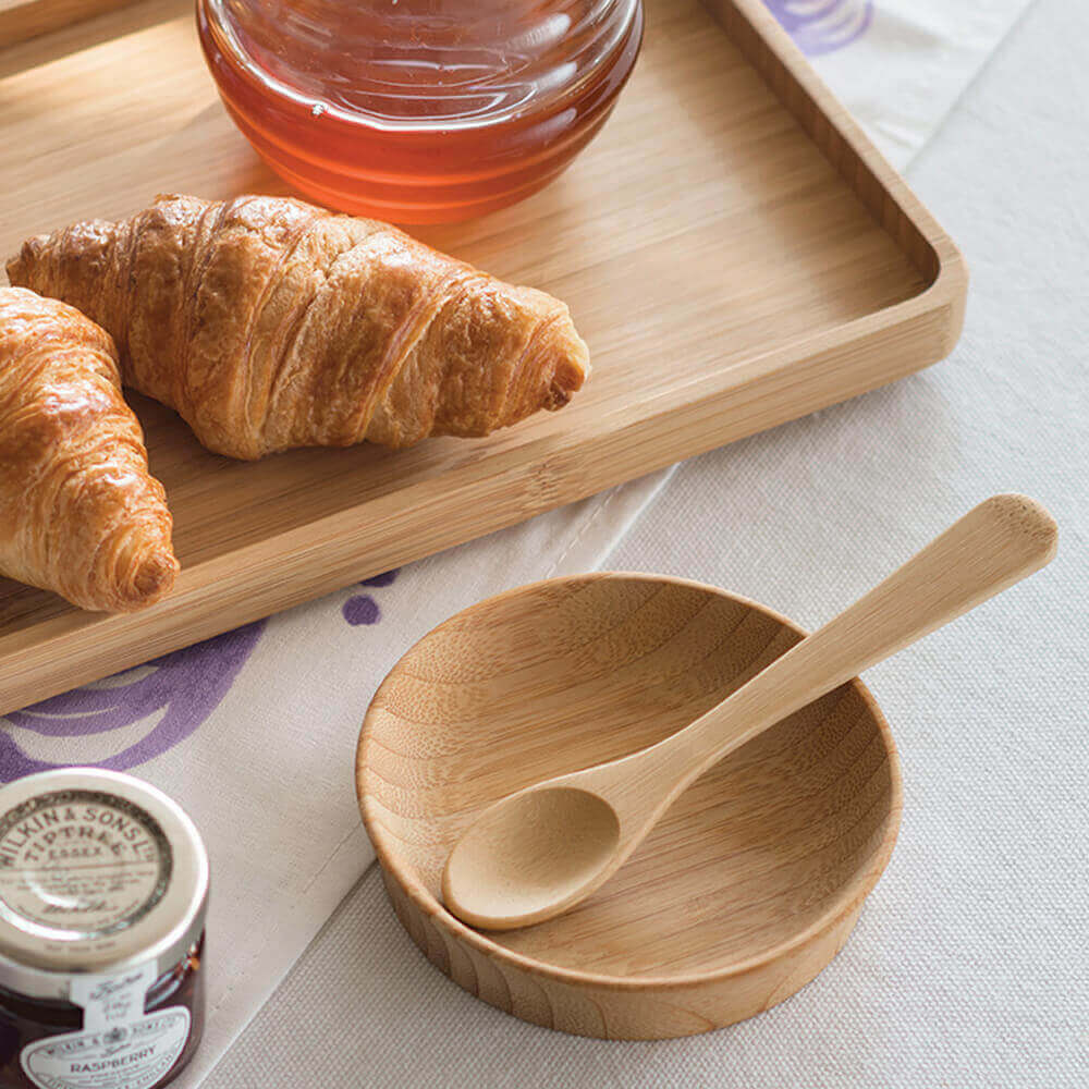 A teaspoon and large condiment cup are next to a bamboo serving tray that holds croissants and a honey pot.