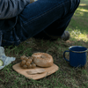 A person in blue jeans sits on a grassy area. A Classic Cutting Bar Board sits next to them. There is a bagel, some grapes, and a spork on the board and a blue enamel mug sits nearby.