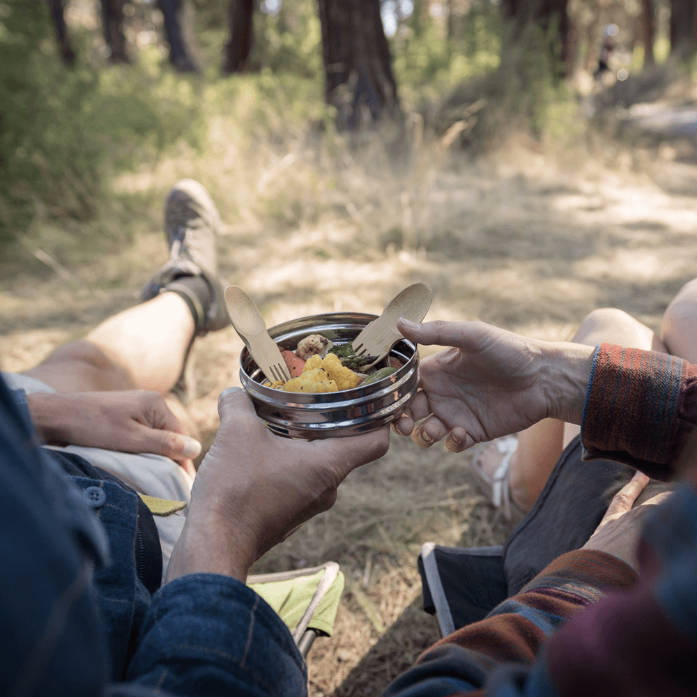 A pair of hikers pause on the trail to eat from a stainless steel container with bamboo sporks.