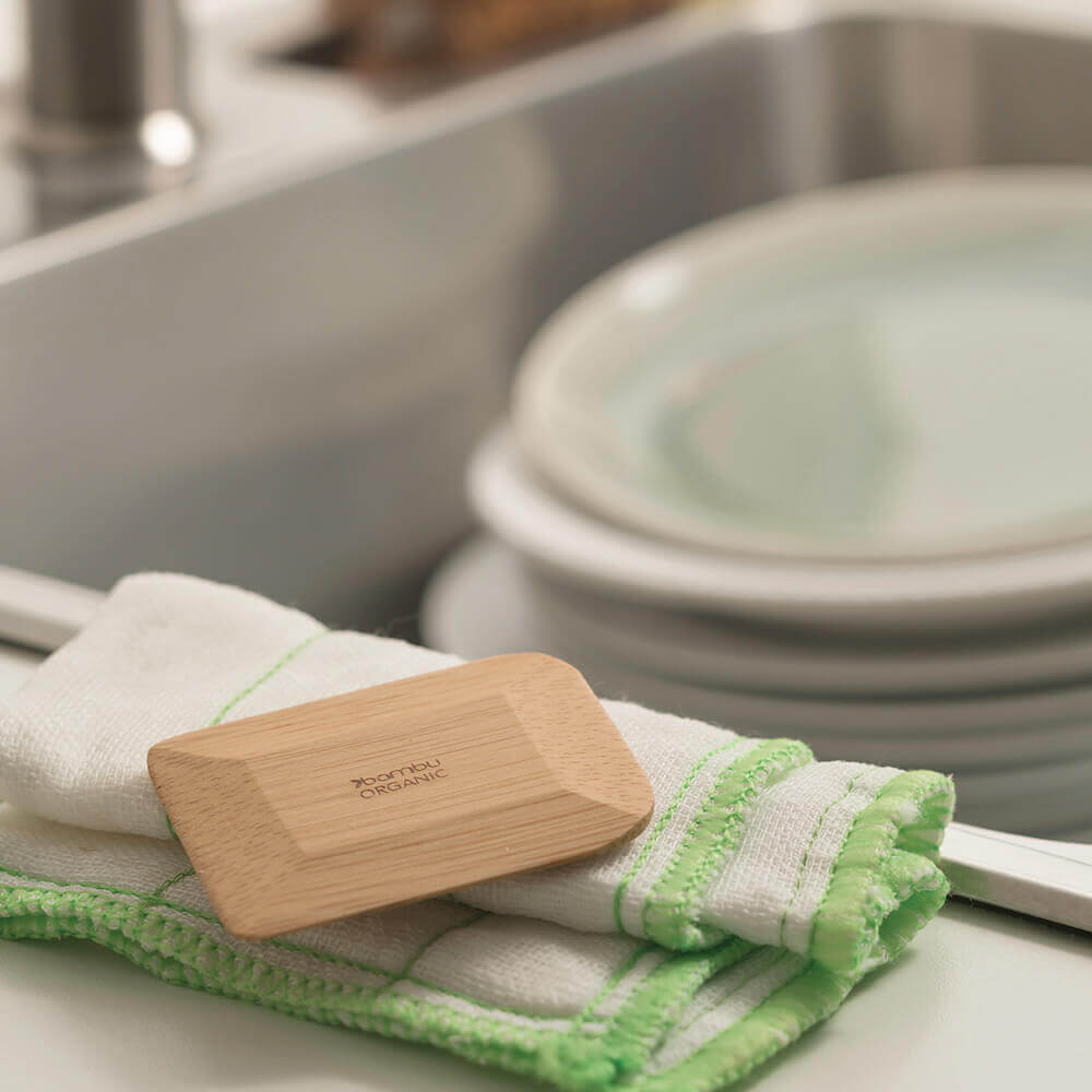 A bamboo pot scraper rests atop a dishcloth next to a sink full of dishes.