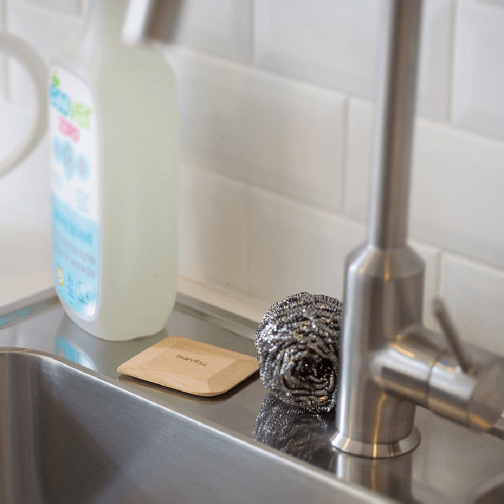 A bamboo pot scraper is resting on a kitchen sink, along with a bottle of dish soap.