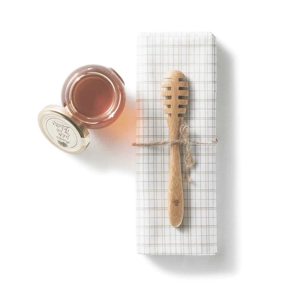 An Organic Bamboo Honey Dipper is resting on a checkered napkin, next to an open jar of honey.