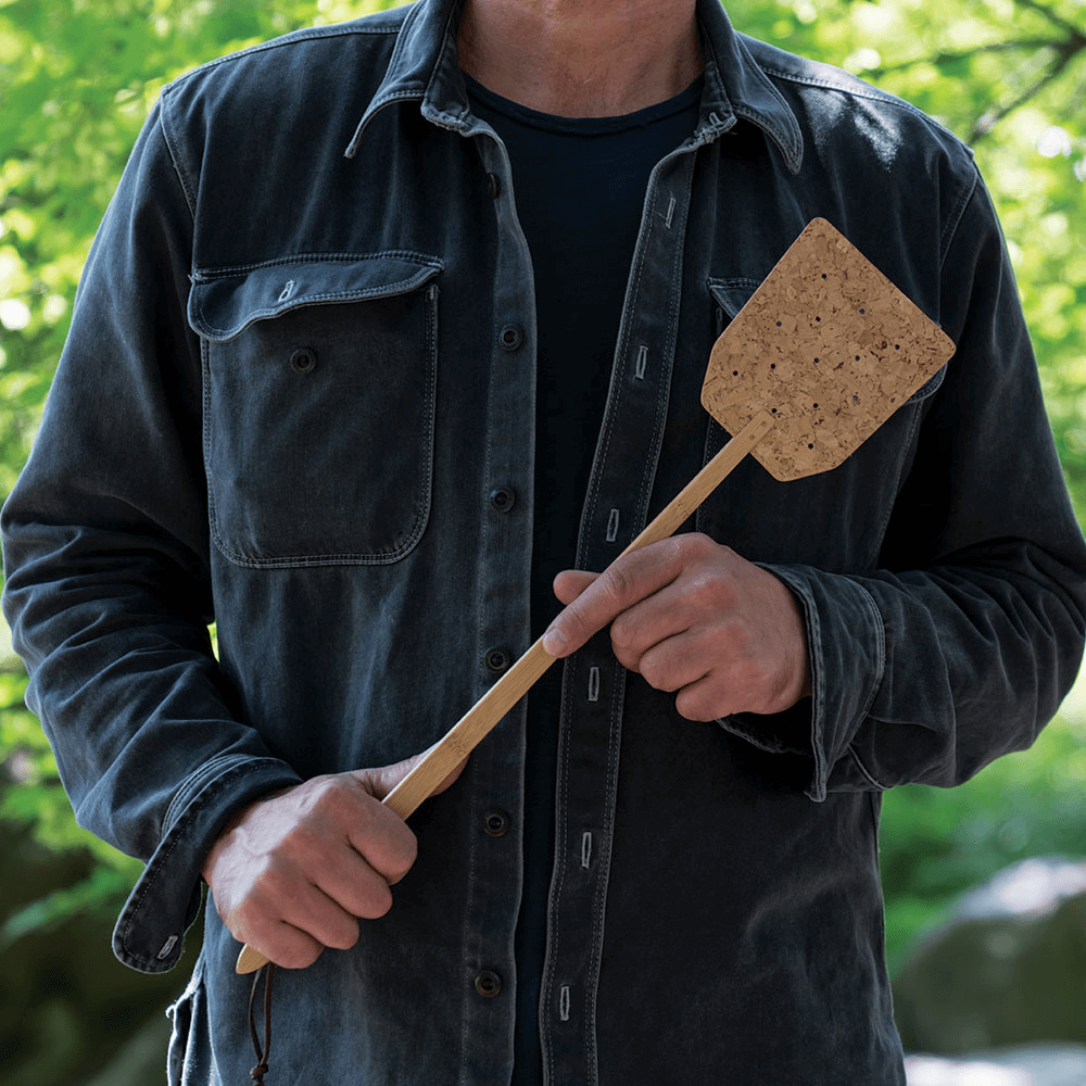 A person wearing a denim shirt is standing outside and holds a Bamboo & Cork Fly Swatter.