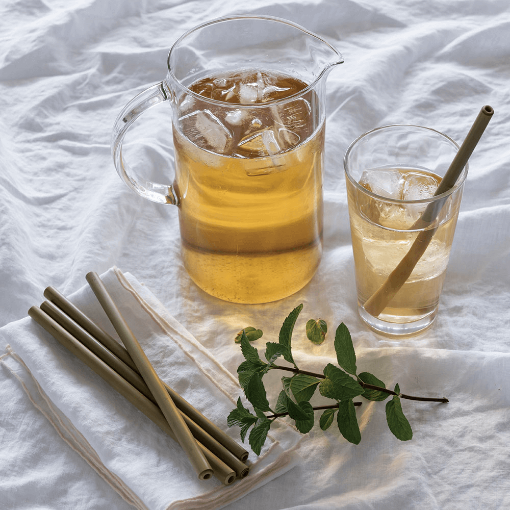 A pitcher of iced tea sits next to a glass that also holds iced tea. A Reusable Bamboo Straw is in the glass, and several more lay on a cloth napkin nearby. 