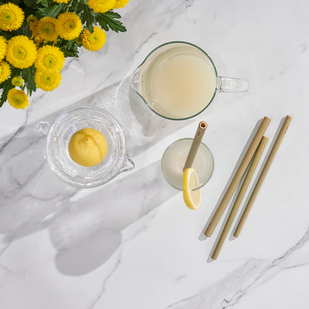 A pitcher of lemonade sits next to several Reusable Bamboo Straws on a marble countertop. A half-squeezed lemon is in a glass dish, and a glass of lemonade has been poured.