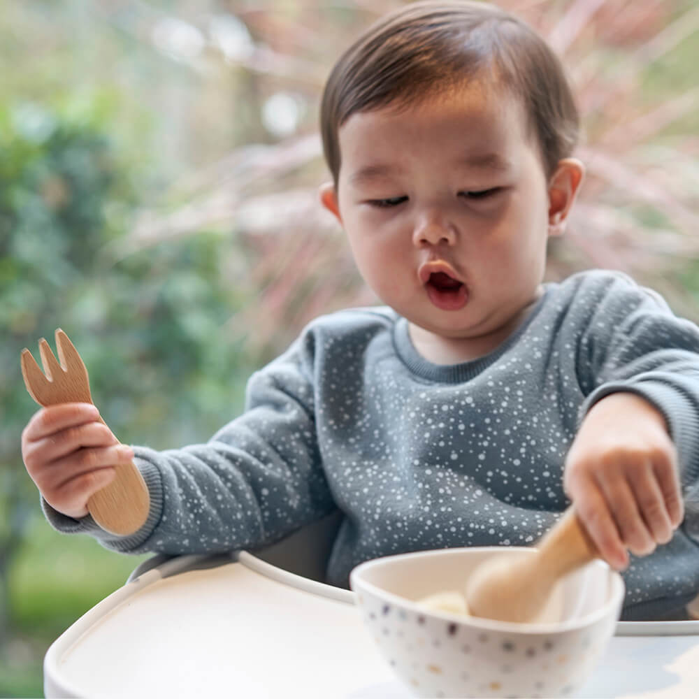 A young child holds a Baby's/Toddler's Fork in one hand, and uses the other to scoop food from a bowl with a Spoon. Designed for children 12M+