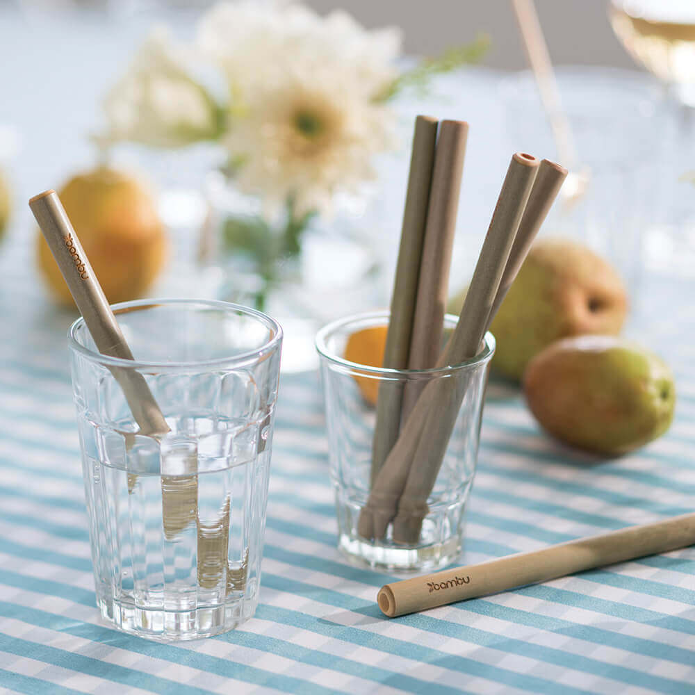A set of Reusable Short Bamboo Straws are displayed in juice glasses on a table set with a blue checkered tablecloth - bambu