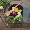 A Bamboo Wok Spatula is laid across the top of a wok filled with colorful vegetables. A pair of tongs are on the counter next to the pan.