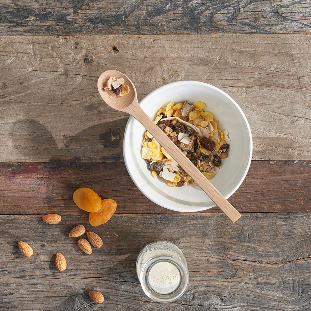 A bamboo trail spoon rests on top os a white ceramic bowl filled with dried fruit and cereal - bambu