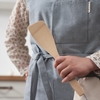 A person in a blue apron holds a bamboo spatula in their left hand.