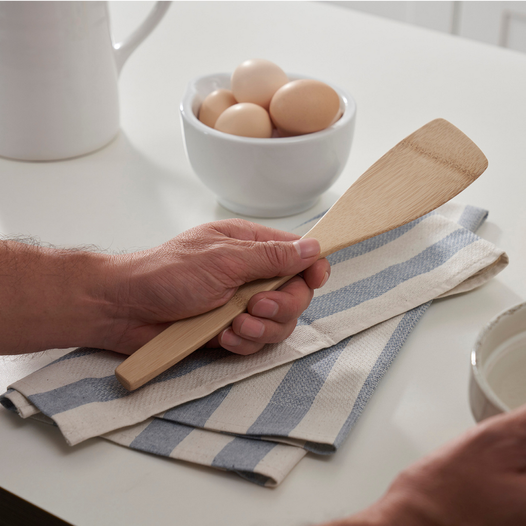 A person holds a left handed spatula while resting their hand on a Meema kitchen towel. A bowl of eggs is nearby on the counter top.
