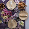 A table is set for three. Each setting has a 9" Veneerware® Fancy Bamboo Plate, a napkin rolled up with a set of Veneerware Cutlery, and water glasses. A flower arrangement is in the middle. A Cork bowl filled with bread rolls is set next to one of the plates. 