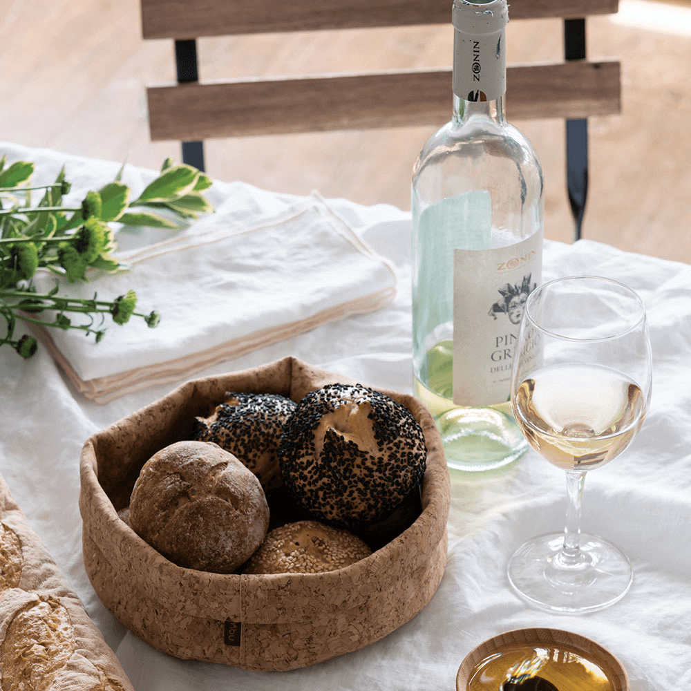 A cork bowl holding bread rolls rests on a table next to a glass of wine. 