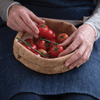 A person wearing a denim apron holds an 8 inch cork bowl full of tomatoes.