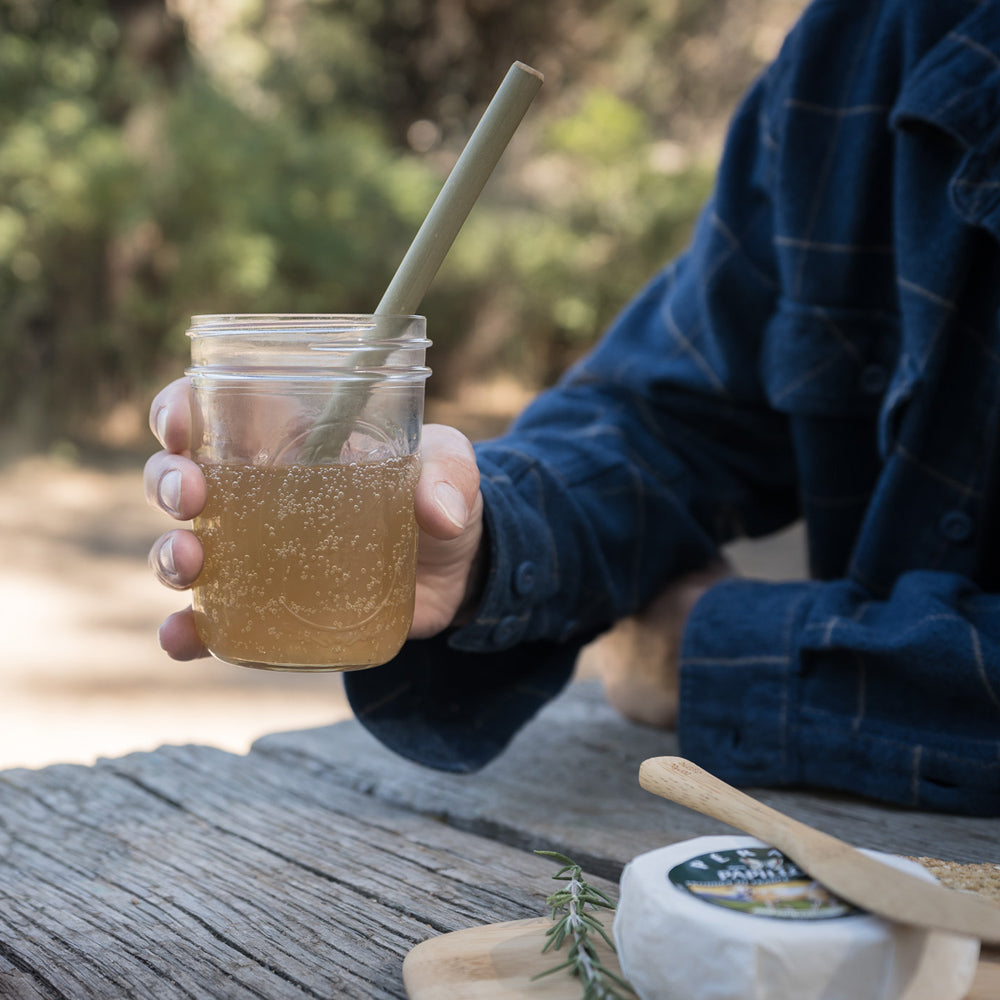 A person in a blue shirt sits at a picnic table. The hold a drinking glass with a Reusable Bamboo Straw in it.