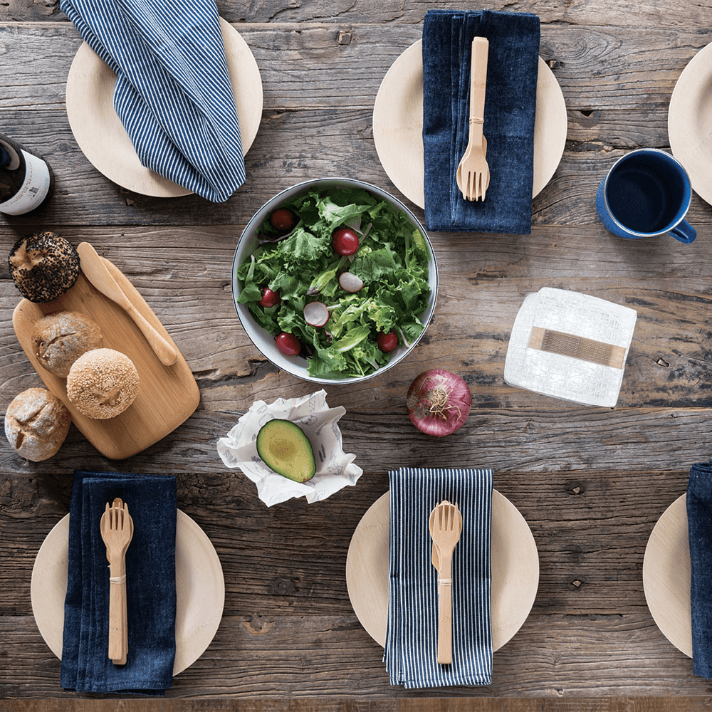 A picnic table is set with 9" Veneerware Round Plates. Each plate has a rustic cloth napkin and a set of reusable bamboo cutlery on top. A bowl of salad and a cutting board with bread on it are in the middle of a table.