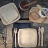 A picnic table place setting holds a 7" square Veneerware plate, a set of reusable bamboo cutlery, and a rustic cloth napkin. A cutting board with bread and cheese is nearby.