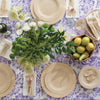 Veneerware® Round Bamboo Plates are stacked on a table that is covered with a purple flower tablecloth. There is a bowl of pears and a vase of flowers on the table.