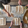 A table is set with 7" and 9" Square Veneerware plates. Several people are reaching into the frame to add beverages, utensils, and napkins to the place settings.