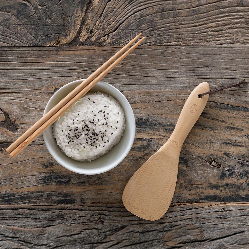 A set of Reusable Bamboo Chopsticks lay across a bowl of rice. A bamboo rice paddle is on the tabletop next to the bowl.