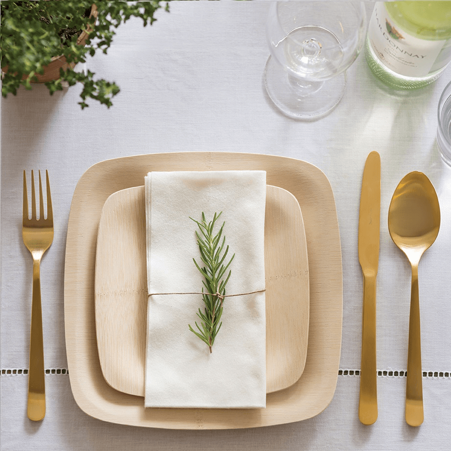 A formal place setting with Veneerware® Square Bamboo Plates is accented with gold flatware and a white linen tablecloth.