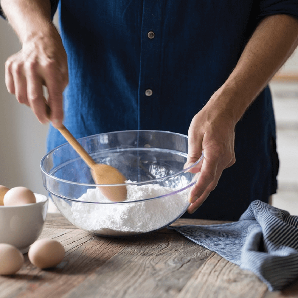 A person in a denim shirt is using a mixing spoon to stir flour in a bowl.