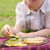 A young child eats from a stainless steel kids tray. There are chips, a dip, and a sandwich on the tray.