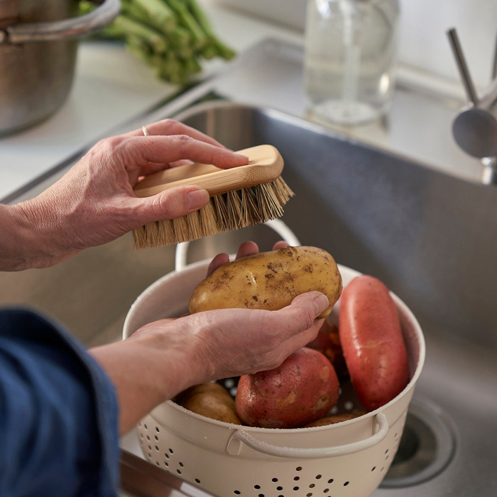 An all-purpose cleaning brush is used to scrub potatoes. The potatoes are in a colander inside a deep sink.