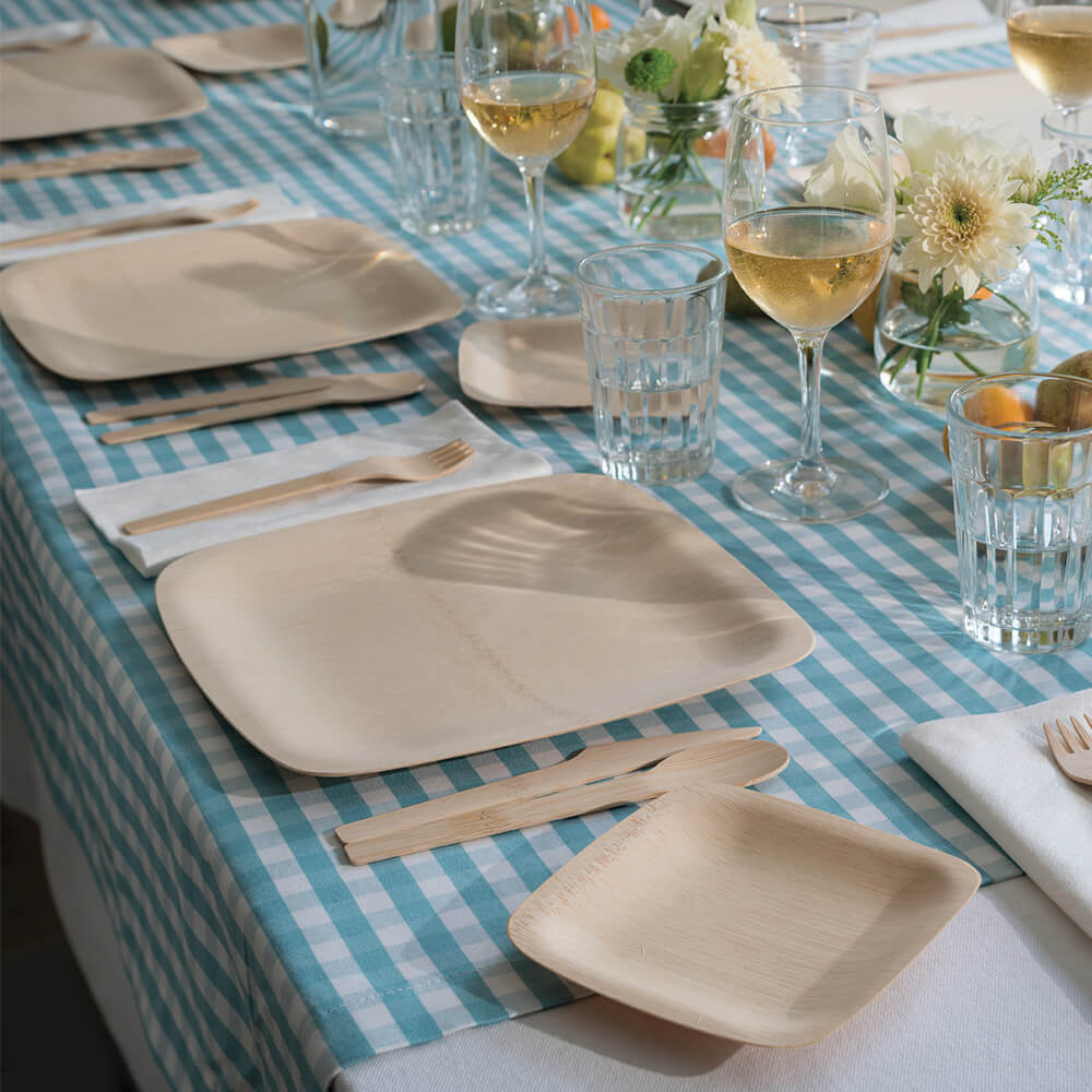 11" Square Veneerware plates are laid out on a table covered with a blue checkered table cloth. Veneerware cutlery is set next to the plates, and there are wine and water glasses above the plates.